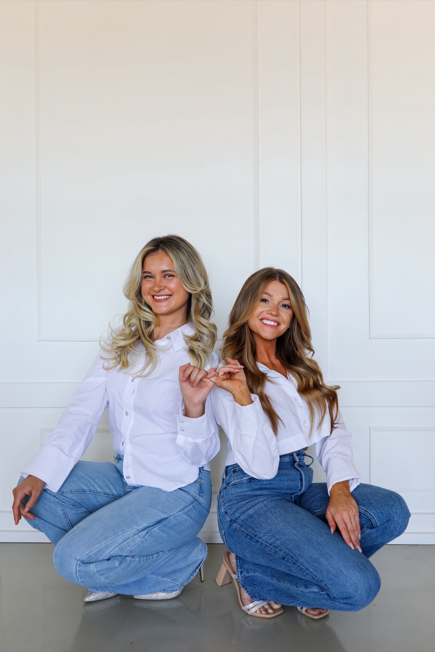 Two women in white shirts and denim crouching against a panelled wall